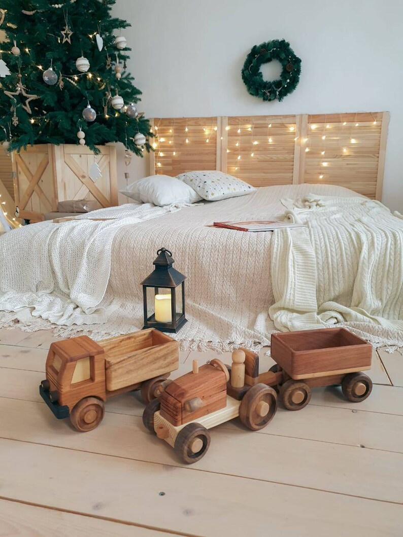 May include: A wooden toy tractor with a trailer, both painted in a natural wood color. The tractor has a small wooden figure in the driver's seat. The toys are on a light wood floor.