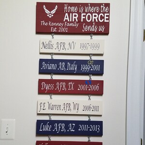 May include: A wooden sign with the text "Home is where the AIR FORCE sends us" and the text "The Kenney Family Est. 2001". The sign is decorated with an Air Force logo and has a red, white, and blue color scheme. Below the main sign are six smaller signs, each with a different Air Force base and the years the family lived there.