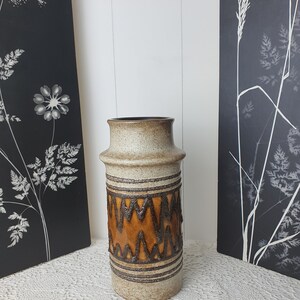 May include: A beige ceramic vase with a brown rim and decorative brown zig-zag pattern. The vase is cylindrical and stands on a white lace tablecloth. Two black and white floral art pieces are in the background.
