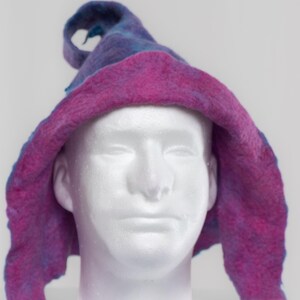May include: A felted hat featuring a pointed, curled top and a wide, floppy brim. The hat displays a mottled blend of deep purple, blue, and vibrant magenta tones, finished with a bright turquoise strap that hangs from the bottom edges.