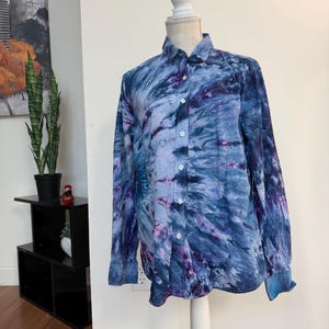 May include: A long-sleeved button-down shirt with a tie-dye pattern in shades of blue and purple. The shirt is displayed on a white mannequin. The shirt has a classic collar and buttoned cuffs.
