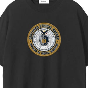 May include: Black t-shirt with a circular graphic featuring the text "CERTIFIED ETHICAL HACKER" and "DIVISION OF DIGITAL WARFARE". The graphic includes a shield, lock, and eagle design in gold, white, and blue.