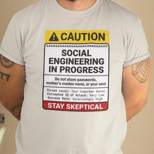 May include: A light gray t-shirt with a warning label design. The label reads "CAUTION SOCIAL ENGINEERING IN PROGRESS" with additional text about passwords and threat levels. The shirt is worn by a person.