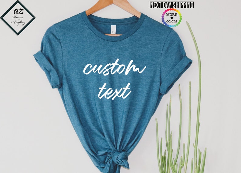 May include: Teal-colored t-shirt with the words "custom text" in white script. The shirt is displayed on a wooden hanger. The sleeves are rolled up. The shirt is knotted at the bottom. The words "Next Day Shipping" are visible.