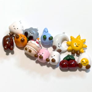 Pode incluir: Uma coleção de pequenos berloques de argila coloridos com vários personagens, incluindo uma nuvem branca, um feijão marrom, uma laranja, uma nuvem cinza, uma gota de chuva azul, um bolo, um marshmallow rosa, um marshmallow branco, uma lua crescente, um sol amarelo, cerejas e um ovo.