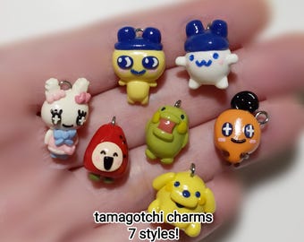 May include: A collection of seven Tamagotchi charms in various styles. The charms are colorful, featuring characters in yellow, white, orange, green, and red. Each charm has a silver loop for attachment. The text "tamagotchi charms 7 styles!" is visible.