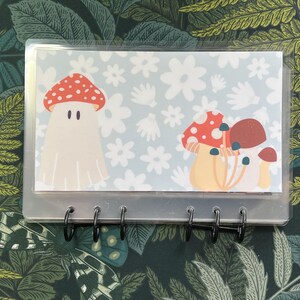 May include: A clear plastic binder with a white background and a colorful illustration of mushrooms. The illustration features a red and white mushroom with a white ghost-like body and a cluster of red and brown mushrooms with white stems. The binder has six silver rings.