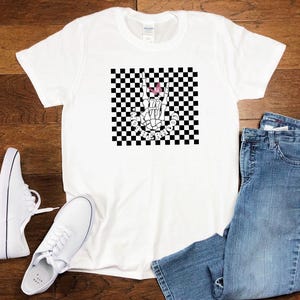 May include: White t-shirt with a black and white checkered pattern and a skeleton hand making the rock and roll sign. The hand has a pink butterfly on it. The text "Rock the Neighborhood" is written below the hand.