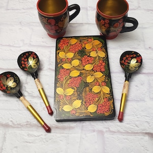 May include: A set of two black wooden mugs with red rims and gold interiors, three black wooden spoons with gold handles and red tips, and a rectangular black wooden tray. All items feature hand-painted floral patterns in red, yellow, and green.