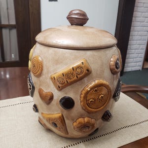 May include: A beige ceramic cookie jar with a removable lid topped by a brown walnut-shaped handle. The exterior is decorated with various raised, three-dimensional cookie shapes in shades of brown, tan, and dark chocolate, featuring textures.