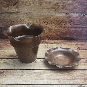 May include: Two copper-colored metal planters with a ruffled edge. The larger planter is taller and has a wider opening. The smaller planter is shallower and has a smaller opening.