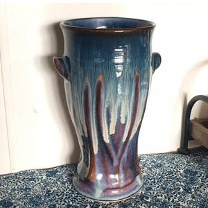 May include: A tall, ceramic vase with a blue and purple glaze. The vase has two small handles and a flared top. The glaze drips down the sides, creating a unique pattern. The vase is a decorative item.