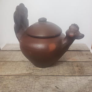 May include: A dark brown ceramic teapot shaped like a bird. The teapot features a rounded body, a lid with a small knob, a tail shaped like a fan, and a head with a beak extending from the opposite side. The surface has a matte finish.