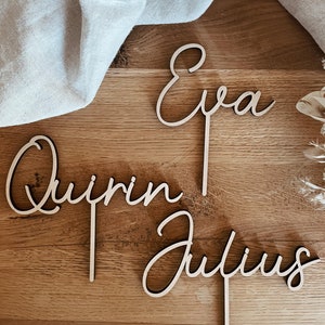 Könnte beinhalten: Drei Holzschilder mit den Namen "Eva", "Quirin" und "Julius" in Kursivschrift. Die Schilder liegen auf einer Holzoberfläche.