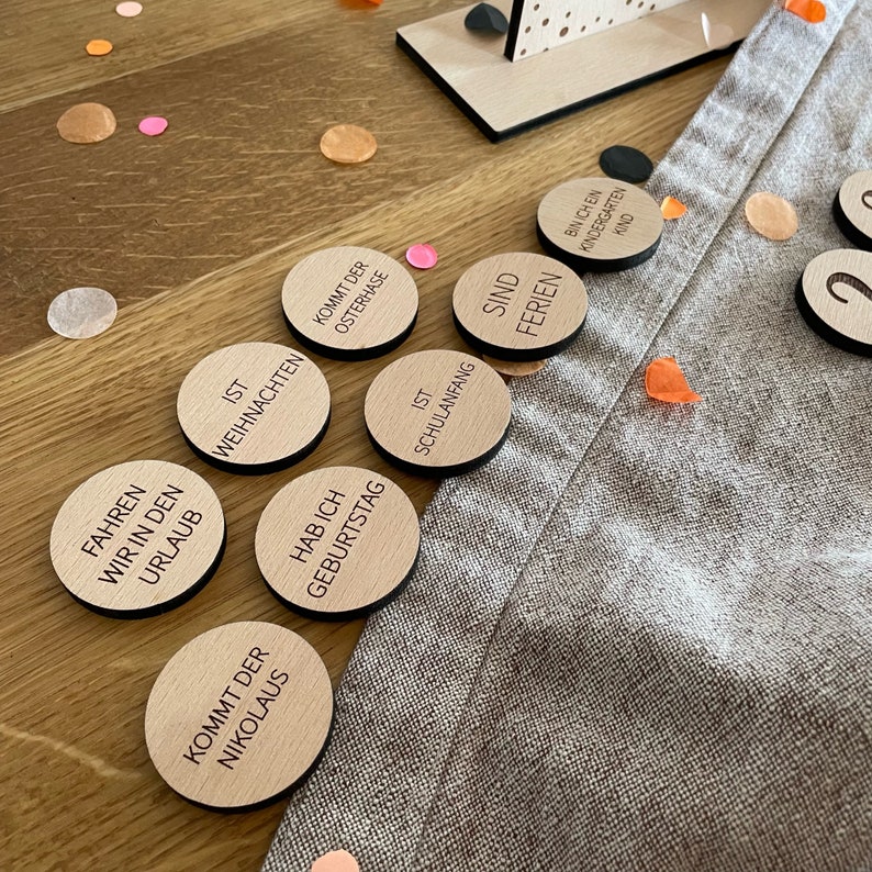 May include: Wooden circle tokens with German text, such as "Ist Weihnachten" (It's Christmas) and "Hab ich Geburtstag" (I have a birthday). The tokens are on a light brown wooden surface with orange and pink confetti.
