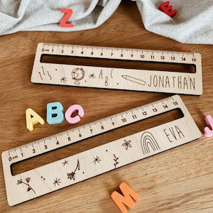 Könnte beinhalten: Zwei Holzlineale, jeweils 15 cm lang, mit eingravierten Markierungen und Namen. Auf einem Lineal steht "Jonathan" und auf dem anderen "Eva". Dekorative Elemente sind Globus, Bleistift und Regenbogen. Bunte ABC-Blöcke und andere Buchstaben sind verstreut.