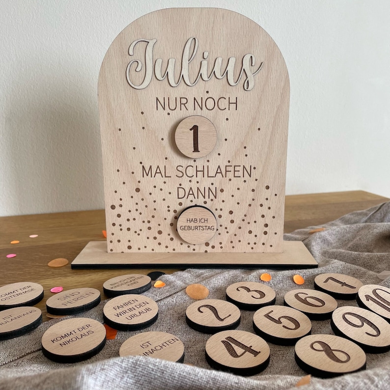 May include: A wooden countdown calendar with a gold glitter background. The calendar says "Julius Nur Noch" and has a number 1 on it. The calendar also says "Mal Schlafen Dann" and "Hab Ich Geburtstag". There are 10 wooden circles with numbers 1 through 10 on them.