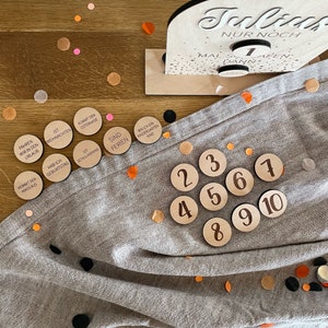 May include: A set of ten wooden circles with numbers 1 through 10 printed on them. The circles are arranged in a semi-circle on a gray fabric surface. There are also several small, colorful confetti pieces scattered around the circles.