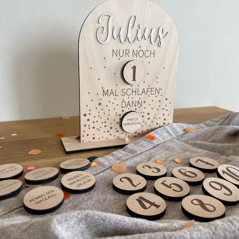 May include: A wooden countdown calendar with the name "Julius" and the text "Nur noch 1 mal schlafen dann hab ich Geburtstag". The calendar has a number 1 on it and a series of wooden circles with numbers 1 through 10 on them.