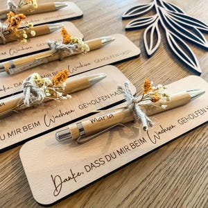 Könnte beinhalten: Mehrere Holzstifte mit silbernen Akzenten und Trockenblumen-Dekorationen werden auf rechteckigen Holzplatten präsentiert. Die Platten weisen eingravierten Text auf, darunter den Namen "Maria". Die Stifte sind mit Bindfäden und kleinen Blumensträußen gebunden.