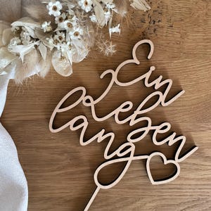 Könnte beinhalten: Holz-Tortenaufsatz mit dem Text "Ein Leben lang" und einem Herz. Der Aufsatz ist hellbraun und hat einen rustikalen Look.