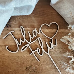 Könnte beinhalten: Holz-Kuchentopper mit den Namen "Julia" und "Felix" in Kursivschrift, mit einem Herzsymbol zwischen den Namen.