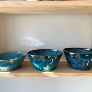 Könnte beinhalten: Drei Keramik-Schalen in verschiedenen Größen, alle mit einem blaugrünen Glazure. Die Schalen sind auf einem Holzregal angeordnet.