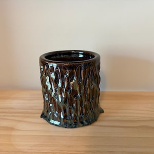 May include: A cylindrical ceramic container with a textured, bark-like exterior. The dark brown glaze gives a glossy finish to the vessel. The container sits on a wooden surface.