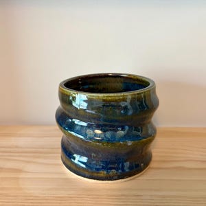 Peut inclure: Un vase en céramique fait à la main avec un design ondulé unique. Le vase présente une finition brillante avec des nuances de bleu, de vert et de marron. L'intérieur est sombre, contrastant avec l'extérieur plus clair. Le vase est posé sur une surface en bois.