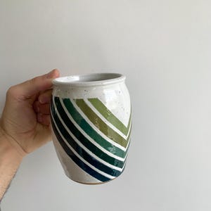 May include: White ceramic mug with a geometric design of green, blue, and brown stripes. The stripes are arranged in a chevron pattern.