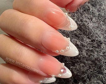 May include: Almond-shaped artificial nails with a nude base and white tips, featuring delicate white line art, small white beads along the smile line, and a single pink rhinestone accent on the ring finger, with the text www.lestarco.com visible.
