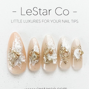 May include: Five nude-colored fake nails with gold and white glitter and embellishments. The nails are arranged in a row on a white background. The text "LeStar Co" and "LITTLE LUXURIES FOR YOUR NAIL TIPS" is above the nails. The text "www.lestar.co.com" is below the nails.