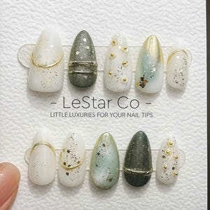 May include: A set of press-on nails in various designs, including white with gold glitter, olive green with silver accents, and gold and white with gold studs. The nails are displayed on a white card with the text "LeStar Co - Little Luxuries For Your Nail Tips."