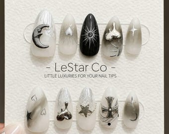 May include: A set of ten almond-shaped press-on nails on a white display card labeled LeStar Co. The nails feature black, white, and gray gradients with silver metallic charms including moons, stars, hearts, and a cross, plus fine line art details.