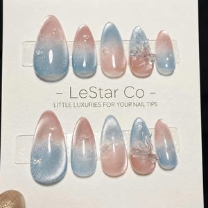 Puede incluir: Un juego de diez uñas postizas en forma de almendra con un degradado rosa y azul y un efecto ojo de gato brillante. Las uñas se presentan sobre una tarjeta blanca con la etiqueta LeStar Co - LITTLE LUXURIES FOR YOUR NAIL TIPS y detalles 3D.