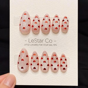 May include: A set of almond-shaped false nails with a glittery pink base and small red heart accents. The nails are arranged in two rows on a white card, with the text "- LeStar Co - LITTLE LUXURIES FOR YOUR NAIL TIPS".
