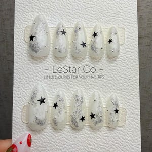 Puede incluir: Un juego de diez uñas postizas en forma de almendra sobre una tarjeta de presentación blanca con la marca LeStar Co. Las uñas son de color blanco roto con detalles de purpurina plateada, estrellas negras y pequeños tachones.