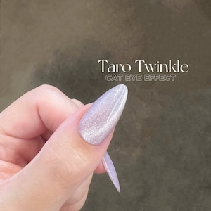 May include: A close-up of a finger with a light purple nail polish with a shimmery finish. The nail polish is called "Taro Twinkle Cat Eye Effect".