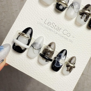 May include: A set of oval-shaped press-on nails in various designs. Some nails are white with black marbling, others are black with white marbling, and some have silver accents. The nails are displayed on a white card with the text "- LeStar Co - LITTLE LUXURIES FOR YOUR NAIL TIPS."