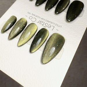 May include: A set of olive green and gold press-on nails with a glossy finish. The nails are arranged on a white card with the text "- LESTAR CO - LITTLE LUXURIES FOR YOUR NAIL TIPS". The nails have a pointed oval shape.