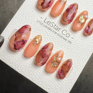 May include: A set of artificial nails with a floral design. The nails are oval-shaped and feature a peach base color with watercolor-style burgundy and brown floral patterns. Some nails have gold accents, glitter, and pearls. The text "- LeStar Co - LITTLE LUXURIES FOR YOUR NAIL TIPS" is visible.