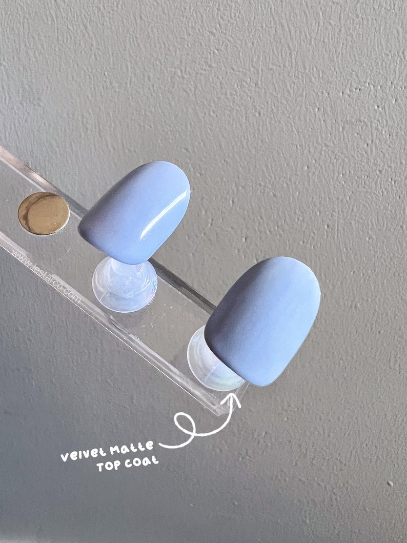 May include: Two light blue nail tips with a velvet matte top coat. The nail tips are displayed on a clear plastic stand.
