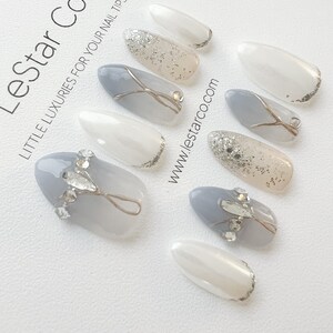 Pode incluir: Conjunto de dez unhas posti&ccedil;as com base branca e glitter prateado, e base cinza com glitter prateado e design de fio prateado. As unhas est&atilde;o dispostas sobre uma superf&iacute;cie branca com o texto "Lestar Co. - Little Luxuries for Your Nail Tips" e "www.lestar.co.com".