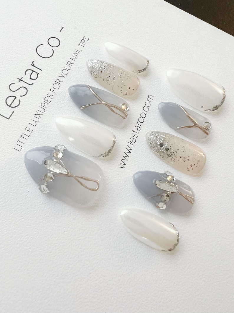 Pode incluir: Conjunto de dez unhas posti&ccedil;as com base branca e glitter prateado, e base cinza com glitter prateado e design de fio prateado. As unhas est&atilde;o dispostas sobre uma superf&iacute;cie branca com o texto "Lestar Co. - Little Luxuries for Your Nail Tips" e "www.lestar.co.com".