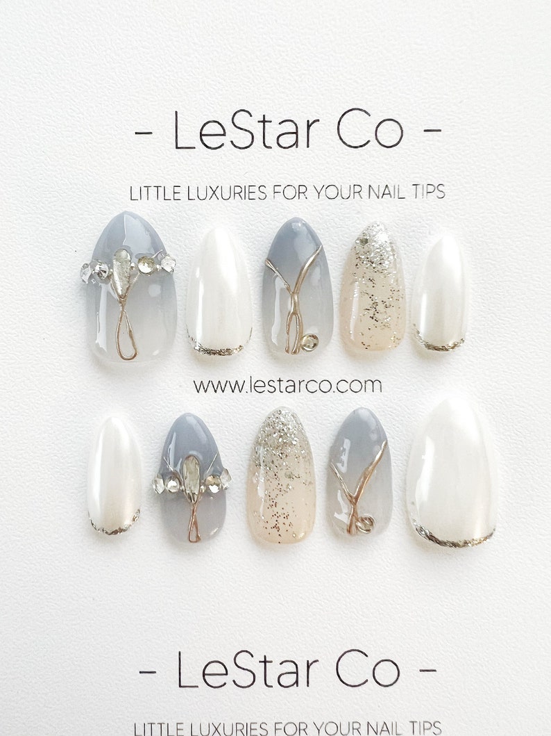 Pode incluir: Um conjunto de dez unhas posti&ccedil;as ovais com diferentes designs. As unhas s&atilde;o brancas, cinzas e bege com detalhes dourados e pedrarias. As unhas est&atilde;o dispostas em duas filas de cinco. O texto "LeStar Co - LITTLE LUXURIES FOR YOUR NAIL TIPS" est&aacute; na parte superior e inferior da imagem. O endere&ccedil;o do site "www.lestarco.com" est&aacute; no meio da imagem.