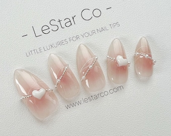 Può includere: Cinque unghie finte a forma di mandorla con un design sfumato rosa e bianco. Ogni unghia è decorata con una fascia color argento e piccole perle bianche. Alcune unghie presentano un piccolo cuore bianco. Il testo "- LeStar Co - LITTLE LUXURIES FOR YOUR NAIL TIPS" è visibile.