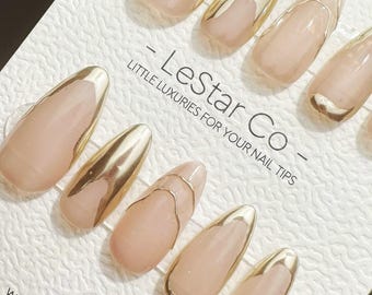 May include: A set of press-on nails with a nude pink base and gold metallic accents. Some nails feature a gold tip, while others have gold designs. The text "- LeStar Co - Little Luxuries for Your Nail Tips" is visible.