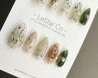 May include: A set of ten almond-shaped press-on nails displayed on a white textured card labeled LeStar Co. The nails feature various designs including olive green, cream, and translucent tones with gold metallic accents, black speckles, and swirls.