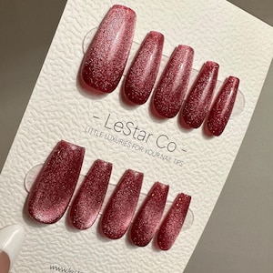 Könnte beinhalten: Ein Set aus glitzernden, roten Kunstnägeln, präsentiert auf einer weißen Karte. Die Karte trägt den Text "- LeStar Co - LITTLE LUXURIES FOR YOUR NAIL TIPS" und eine Website-Adresse. Die Nägel sind tiefrot.