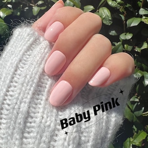 Op de afbeelding: Close-up van een hand met vijf nagels die in een lichtroze kleur zijn gelakt. De nagels zijn kort en ovaalvormig. De hand rust op een witte gebreide stof. De tekst "Baby Pink" is in zwarte letters op de stof geschreven.